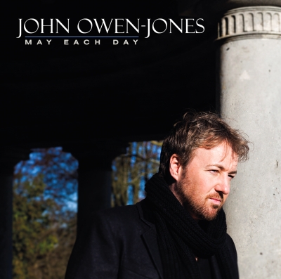 A bearded man in a dark coat and scarf stands beside a pale stone column, with trees visible through dark openings behind him.